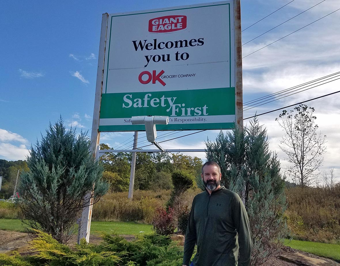 Dan in front of OK Grocery welcome sign